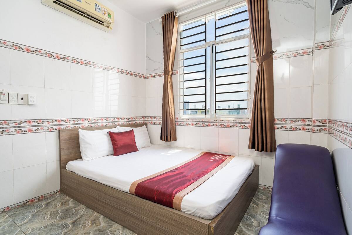 A hotel room with a bed, a window with curtains, a chair, and a wall-mounted air conditioner.