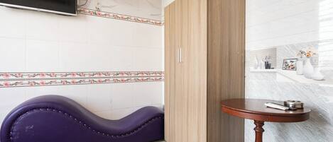 A modern bathroom with a wooden wardrobe, a purple cushioned bench, a round wooden table, and a wall-mounted fan.