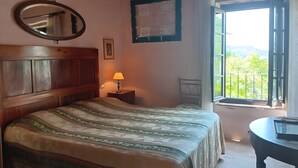 A bedroom with a wooden headboard, a bed with patterned bedding, a bedside lamp, a wall-mounted mirror, a framed picture, and a window with a view of greenery. - Villa 'Olivi' with Shared Pool, Private Garden and Wi-Fi (Nocchi)
