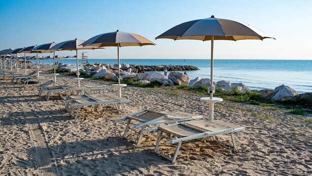 Ein Strand mit Liegestühlen, Sonnenschirmen, felsigem Ufer und ruhiger See.