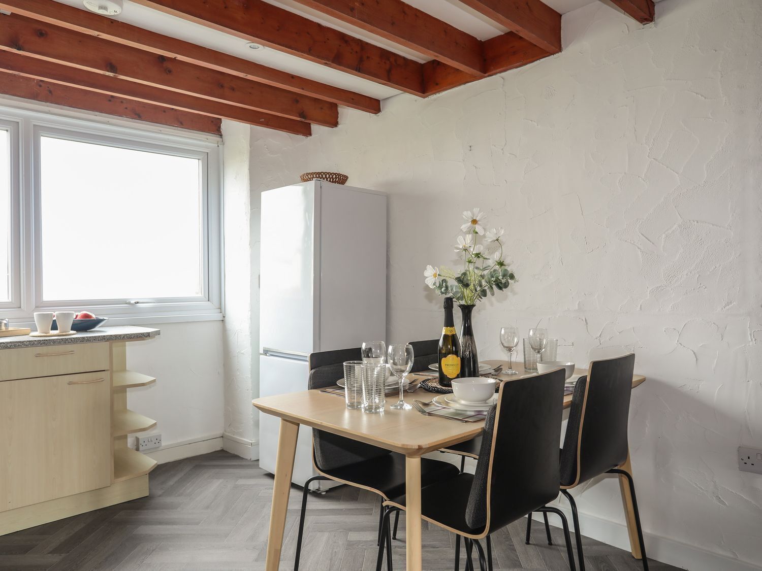 Uma sala de jantar com uma mesa de madeira posta para quatro pessoas, uma geladeira branca e uma janela com luz natural.