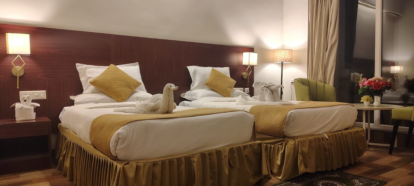 Habitación de hotel con dos camas, un sillón verde, una mesita con un florero y una ventana con cortinas.