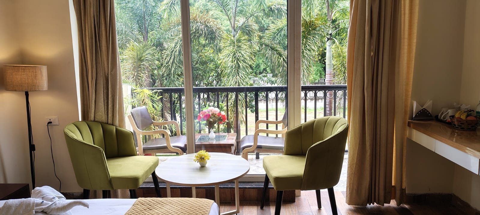 A room with two green chairs around a small round table, a bed, and a view of a balcony with a railing and greenery outside.