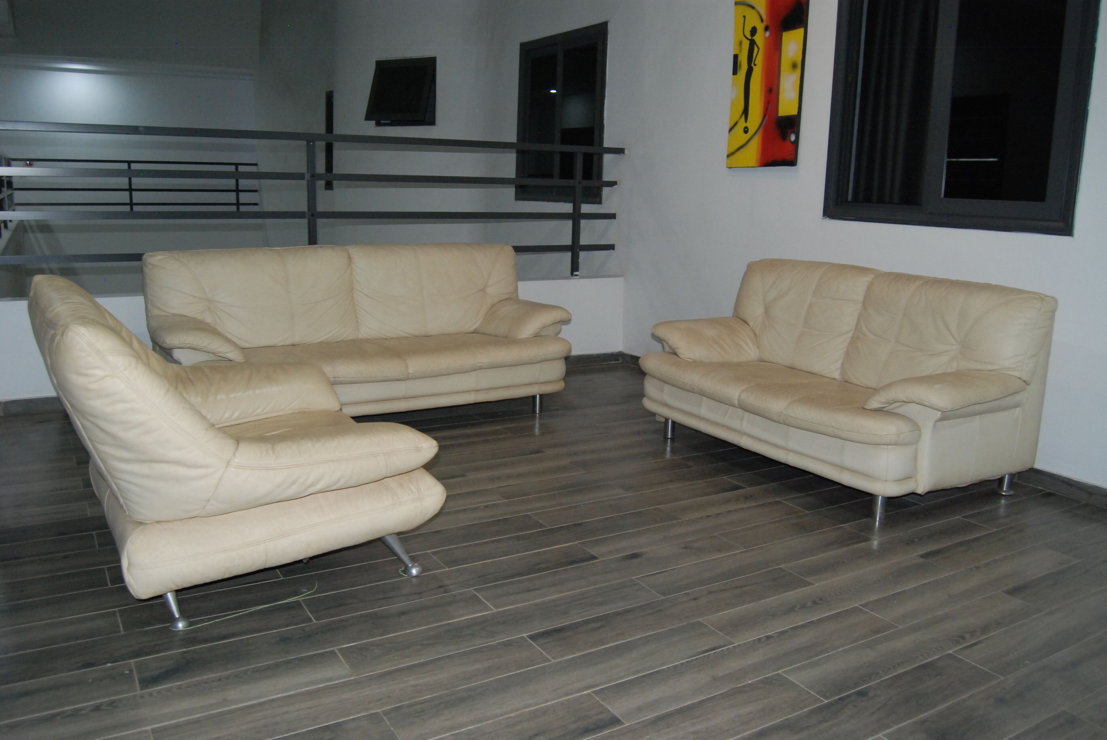 A modern living room with two beige leather sofas, a dark wood floor, a staircase with metal railings, and a painting on the wall.