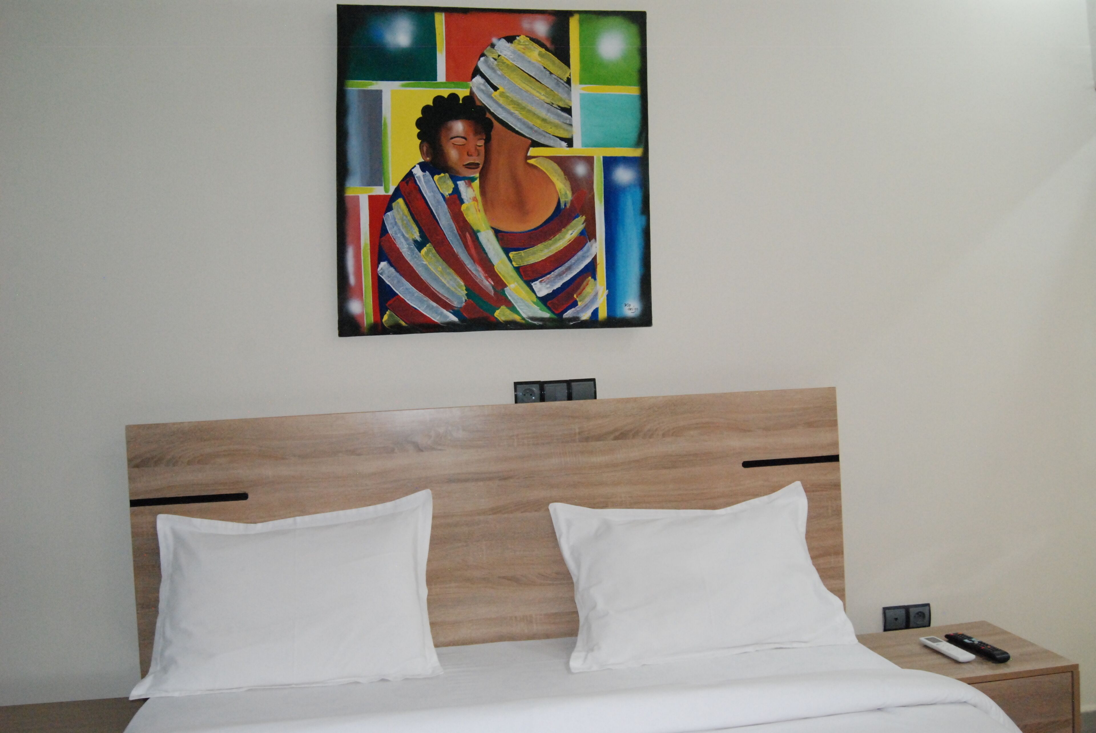 Habitación de hotel con cabecera de madera, dos almohadas blancas, una cama, una mesita de noche con un control remoto y un cuadro en la pared.