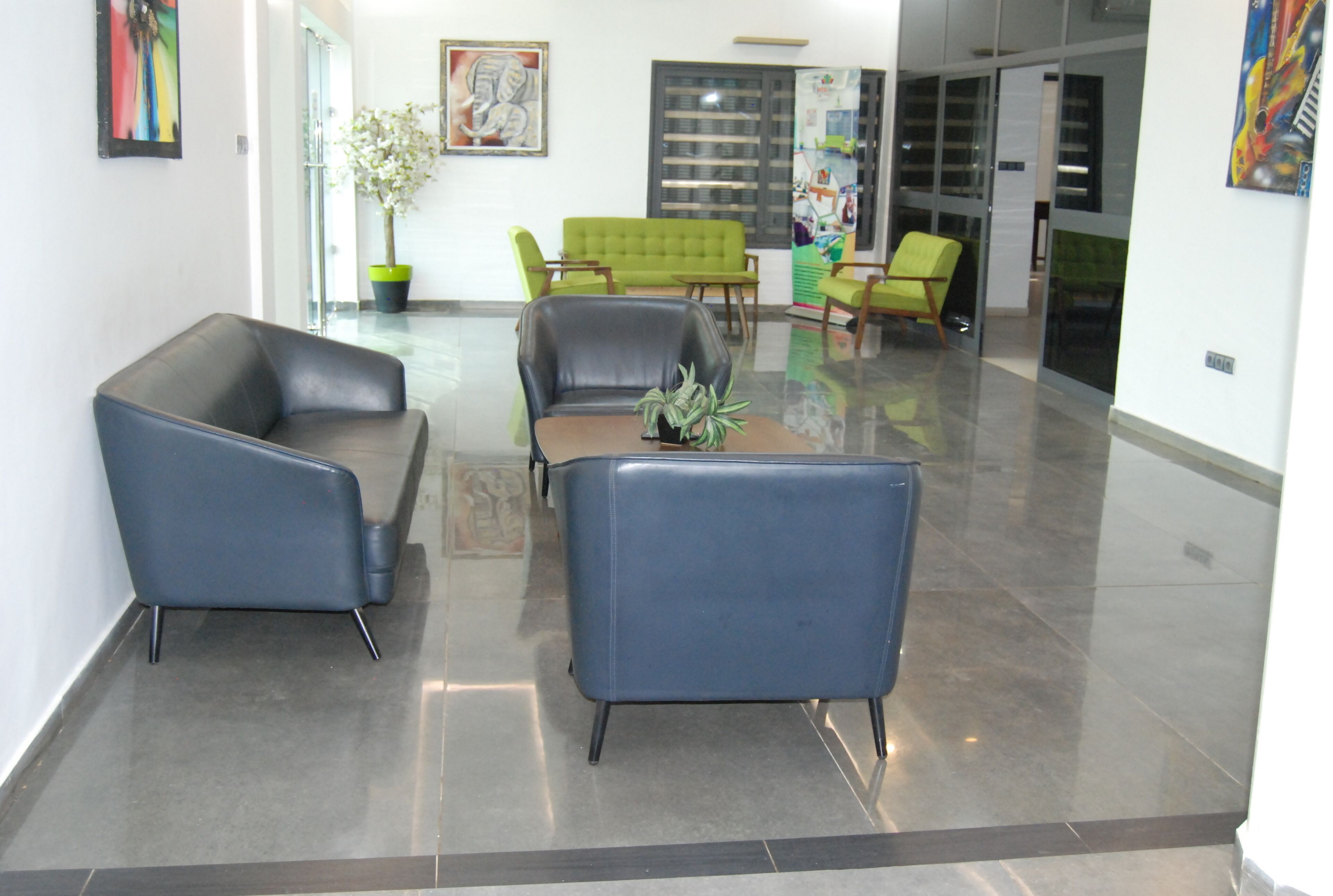 A modern lobby with a blue sofa, a green sofa, and a small table with a plant. There are potted plants and artwork on the walls.