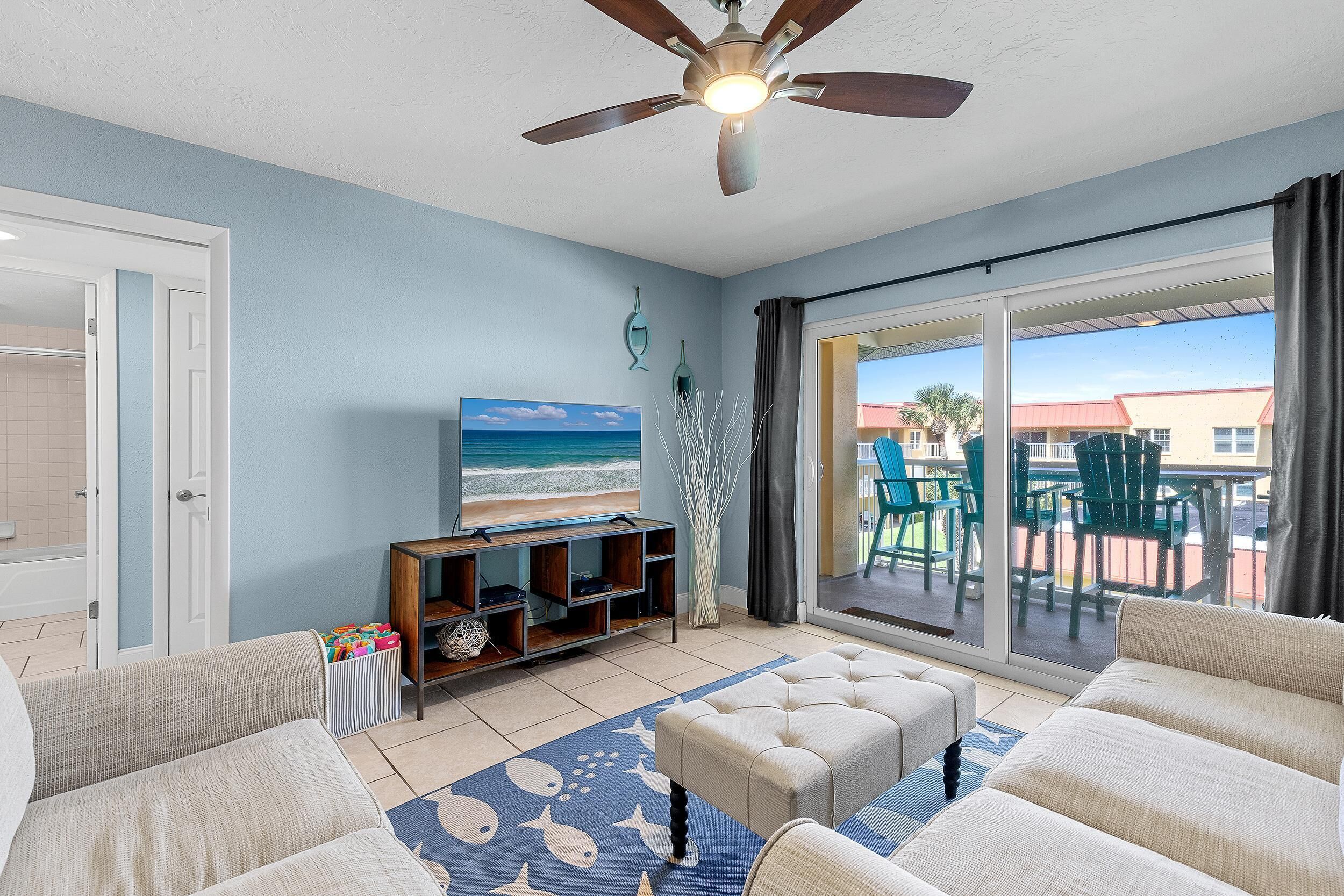 A living room with a ceiling fan, a television, and a view of the beach through large sliding glass doors.
