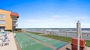 A shuffleboard court adjacent to a poolside area with lounge chairs and a building with a red roof. - Point East 320 (New Smyrna Beach)