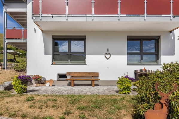 A building with a white facade, two windows with blinds, a wooden bench, and a heart-shaped decoration on the wall. - Apartment 'Holzwieb' with Private Terrace, Shared Garden and Wi-Fi (Unterkirnach)