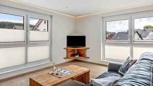 A modern living room with large windows, a flat-screen TV, a wooden coffee table, and a grey sofa. - Apartment 'Holzwieb' with Private Terrace, Shared Garden and Wi-Fi (Unterkirnach)
