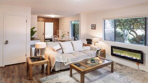Una sala de estar moderna con sofá, mesa de centro y chimenea. Se observa un comedor con sillas y una mesa al fondo.