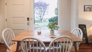 Un comedor con una mesa de madera, sillas blancas, una planta en maceta y vistas a árboles a través de un ventanal grande.