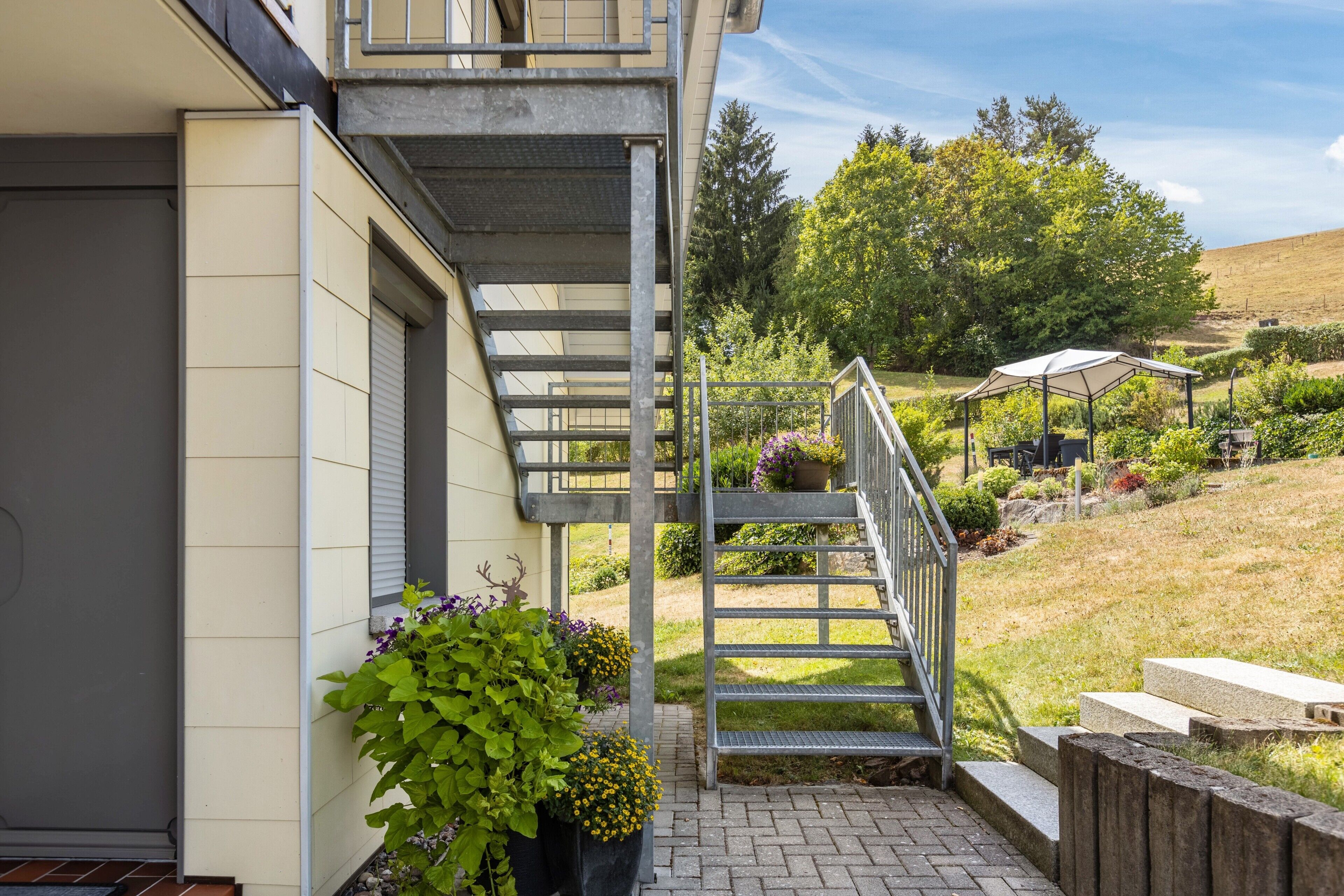 Propiedad residencial con una escalera que lleva a un balcón, rodeada de vegetación y un jardín cercado.