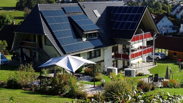 Une maison moderne dotée de panneaux solaires sur le toit, d’un espace de détente extérieur abrité et offrant une vue sur un quartier résidentiel.
