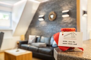 Un jouet en peluche est posé sur un comptoir, accompagné d’une pancarte sur laquelle on peut lire « Schwarzwaldtraum » et « Schön, dass Sie unsere Gäste sind ! ». La scène se déroule dans le hall d’un hôtel, où l’on aperçoit un canapé de couleur sombre et une petite table.
