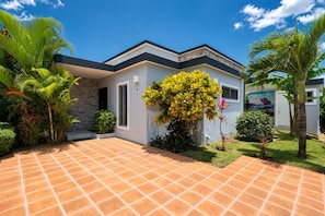 A modern house with a tiled patio, surrounded by tropical plants and a clear blue sky.