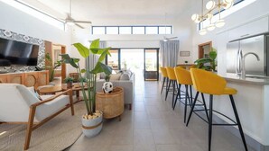 Una sala de estar moderna con un sofá, una mesa de centro y sillas, una cocina con refrigerador y taburetes de bar, y ventanas grandes.