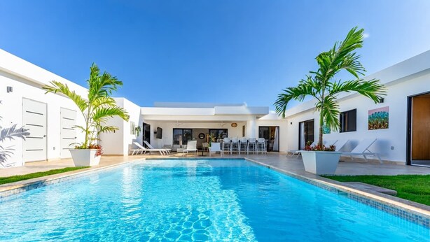 Un área de piscina moderna con agua azul cristalina, paredes blancas y palmeras.