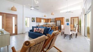 A spacious living area with a sofa, dining table, and chairs, a kitchen in the background, and large windows. - Luxurious 4-bedroom villa in Casa Linda. (Sosúa)