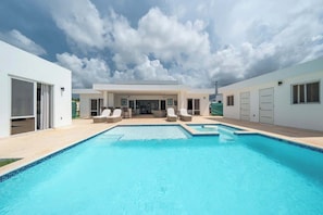 Un área de piscina moderna con agua azul cristalina, una zona de estar techada y un edificio blanco con ventanas y puertas amplias.