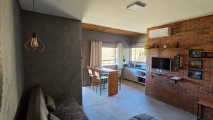 A modern living room with a brick accent wall, a dining area, and a kitchen with a TV and wine glasses. - Villa da Serra Studio Ibitipoca (Lima Duarte)