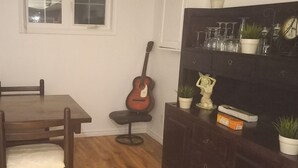 A dining area with a wooden table and chairs, a guitar leaning against the wall, and a shelf with glassware and plants. - Walk out to Salmon Creek (Bowmanville)