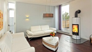 Un salon moderne avec un canapé blanc, une table basse ronde ornée d’un vase de fleurs, un luminaire fixé au mur et un poêle à bois.
