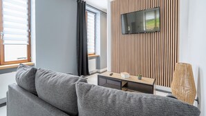 A modern living room with a grey sofa, a wooden TV stand, and a large window with blinds.