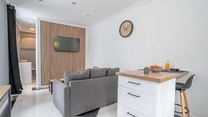 A modern living room with a grey sofa, a small kitchen area with a wooden countertop, and a wall-mounted TV.