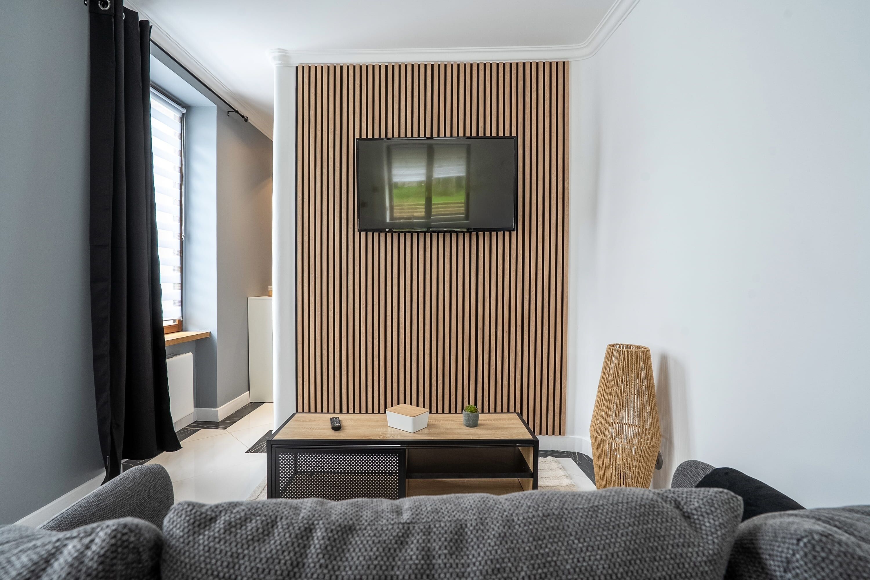 Una camera moderna con una TV montata su un pannello di legno, un tavolino con una scatola e una pianta, e una lampada grande intrecciata.