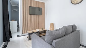 A modern living room with a grey sofa, a wooden slatted wall, a flat-screen TV, and a large wall clock.