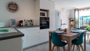A modern kitchen with white cabinets, a wooden dining table with teal chairs, a clock on the wall, and a view of the outdoors through a glass door.