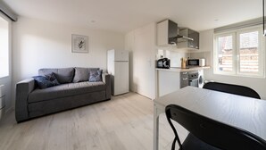 A compact living space with a grey sofa, a dining table, and a kitchen area including a microwave and a washing machine. - Le Nid du Beffroi - Studio centre Doullens (Doullens)