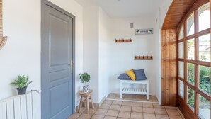 A hallway with a grey door, a small stool with a plant, a white bench with cushions, and a wooden window frame. - Le gite de la petite famille - proche lacs (Cornod)