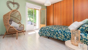 A bedroom with a bed, a wicker chair, a wooden wardrobe, and a sliding glass door leading to a balcony. - Le gite de la petite famille - proche lacs (Cornod)