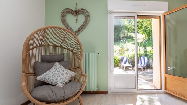 A wicker chair with cushions, a heart-shaped decoration on the wall, and a sliding glass door leading to an outdoor area with chairs. - Le gite de la petite famille - proche lacs (Cornod)