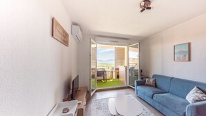A modern living room with a sofa, a small table, and a TV. There is a sliding glass door leading to an outdoor area with a patio and a view of the mountains.