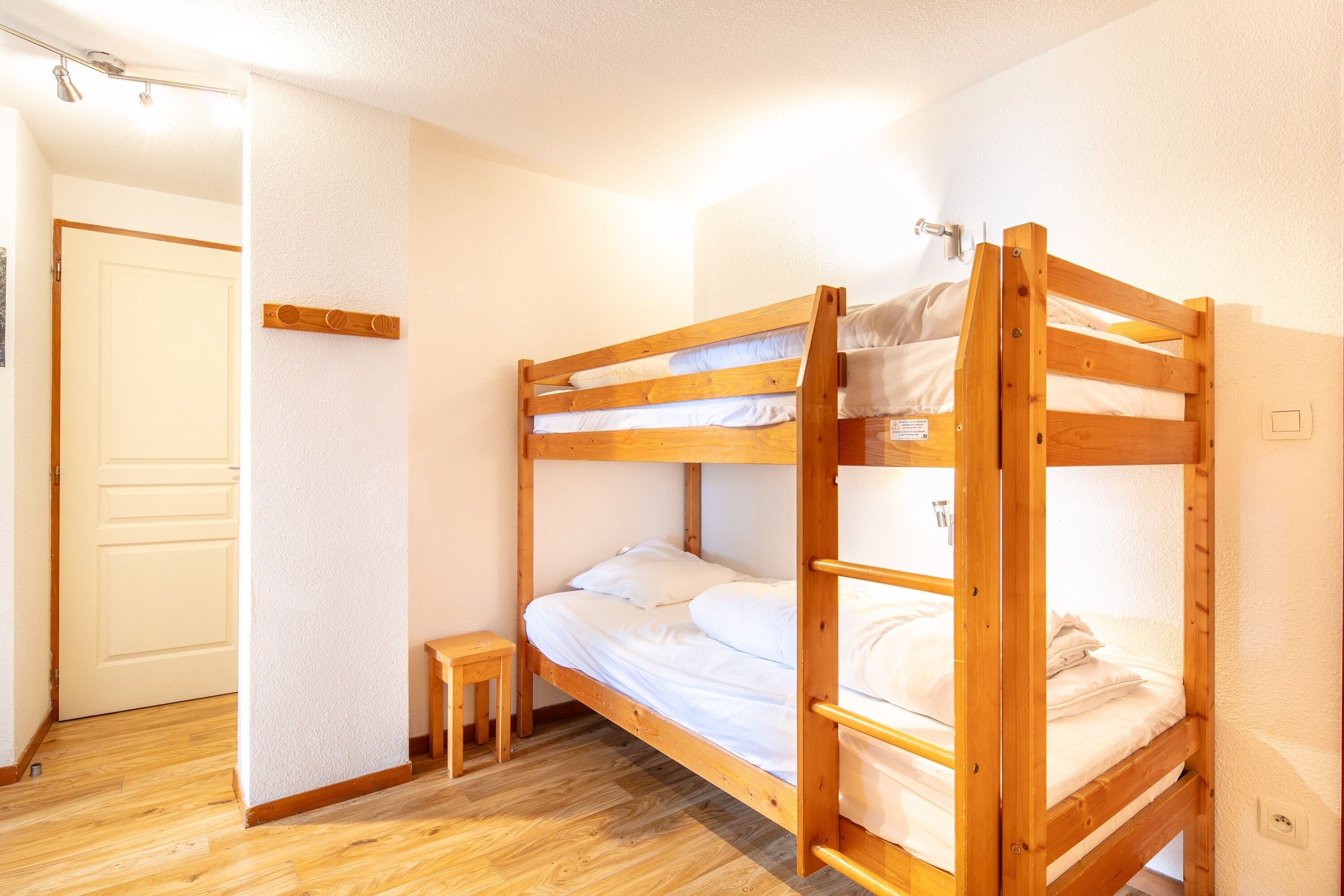 A bunk bed room with a wooden frame, white bedding, and a small wooden stool.