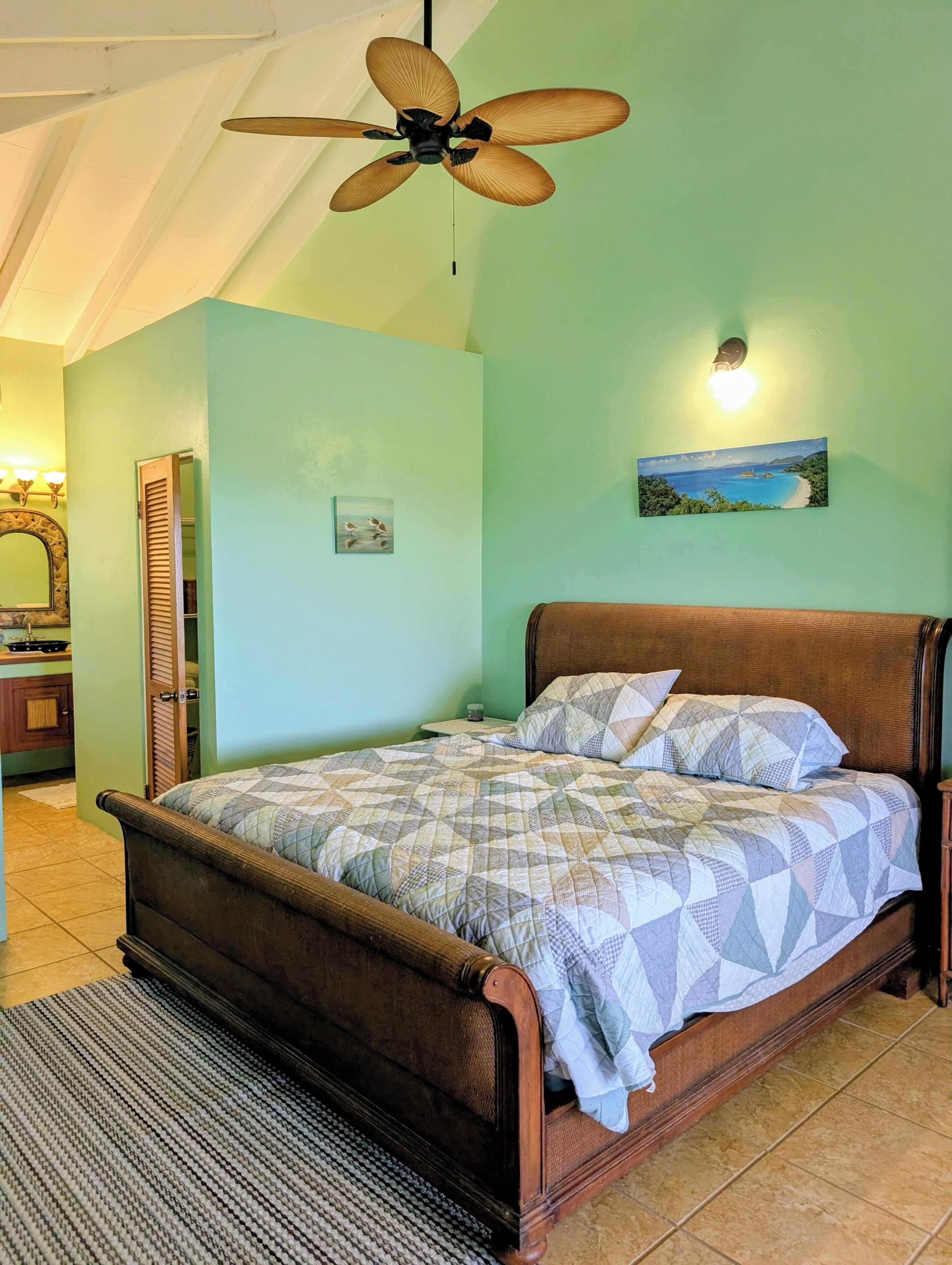 A bedroom with a bed, a ceiling fan, a painting, and a bathroom visible through an open door.