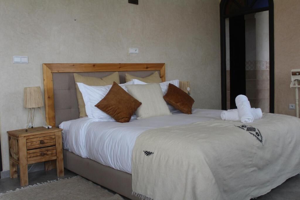 Un dormitorio con cabecera de madera, cama con ropa de cama blanca y marrón, una mesita de noche de madera y una toalla blanca sobre la cama.