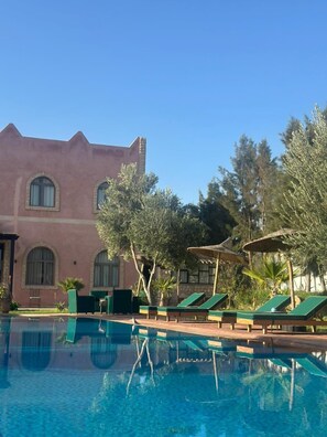 A swimming pool with lounge chairs, a building with arched windows, and surrounding trees.