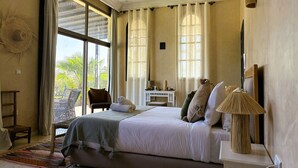 A bedroom with a large bed, a bedside table with a lamp, a chair, and a view of the outdoors through a window. - Dar mimosa (Sidi Kaouki)