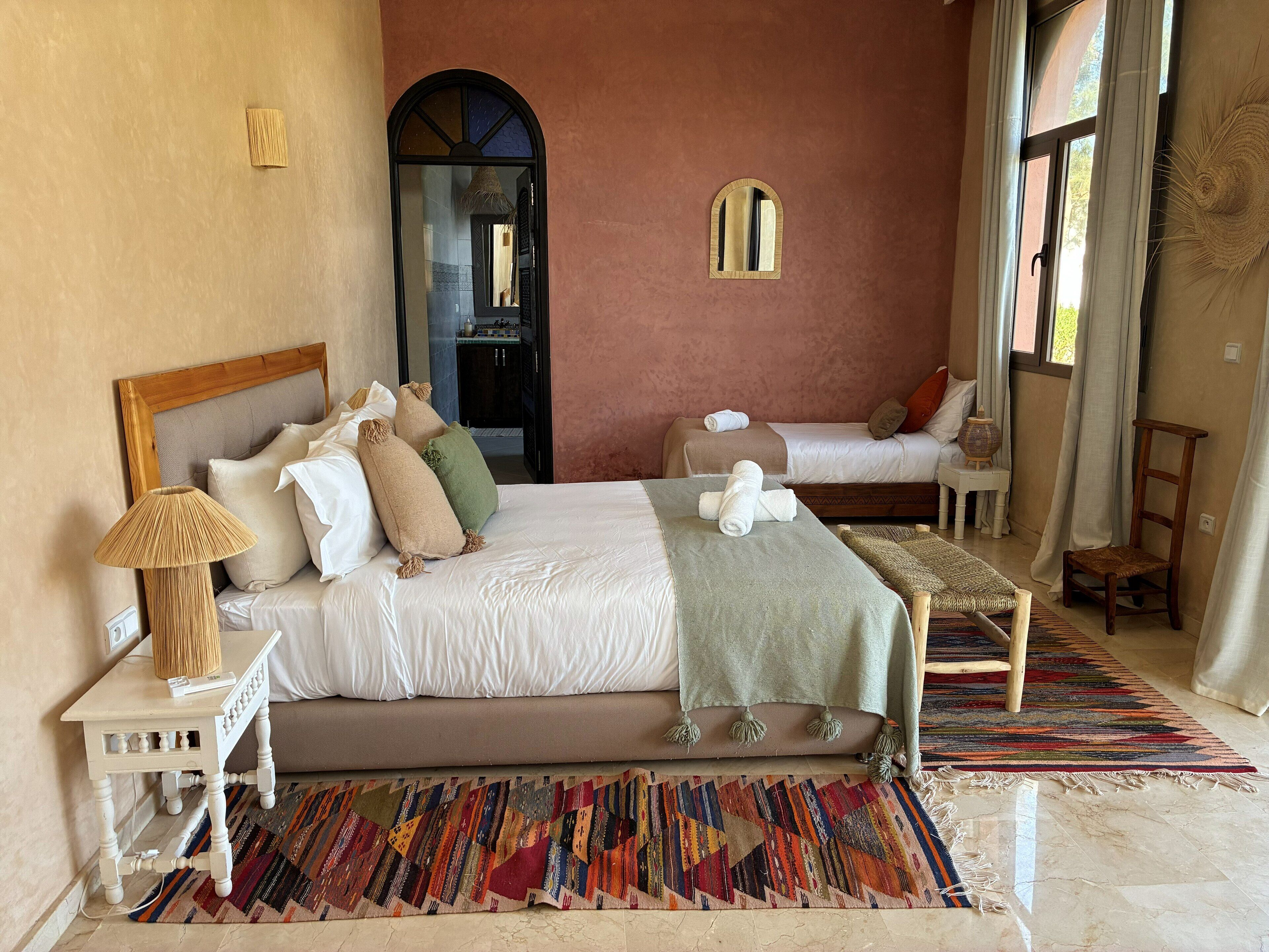 A spacious bedroom with a large bed, a smaller bed, a nightstand, a chair, a colorful rug, and a mirror on the wall.