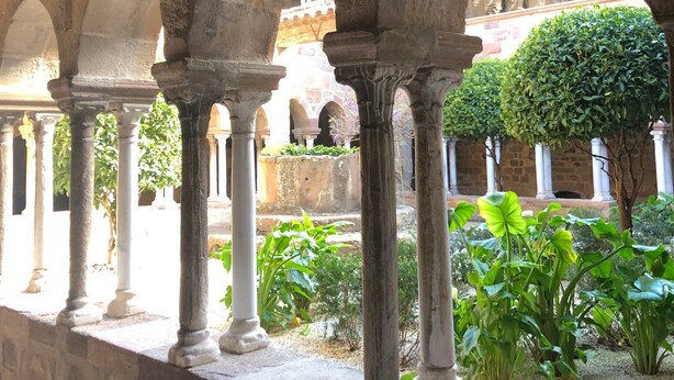 An ancient stone structure with arches and columns, a garden with plants, and a brick building in the background.