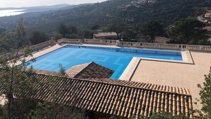 A swimming pool with a tiled deck, surrounded by a fence, overlooking a hilly landscape with buildings.