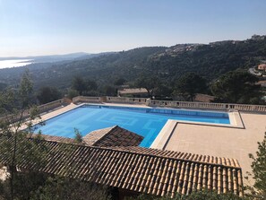 A swimming pool with a tiled deck, surrounded by a fence, overlooking a hilly landscape with buildings. - Apartment 'Chez Skadi' with Shared Pool and Wi-Fi (Roquebrune-sur-Argens)