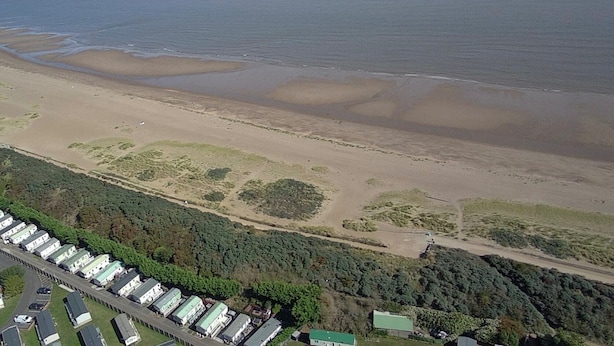 An aerial view of a coastal area with a sandy beach, a dense forested area, and a row of white buildings with green roofs. - 2 Bedroom Caravan Sleeps 6 -free Parking -Tv -wifi (Skegness)