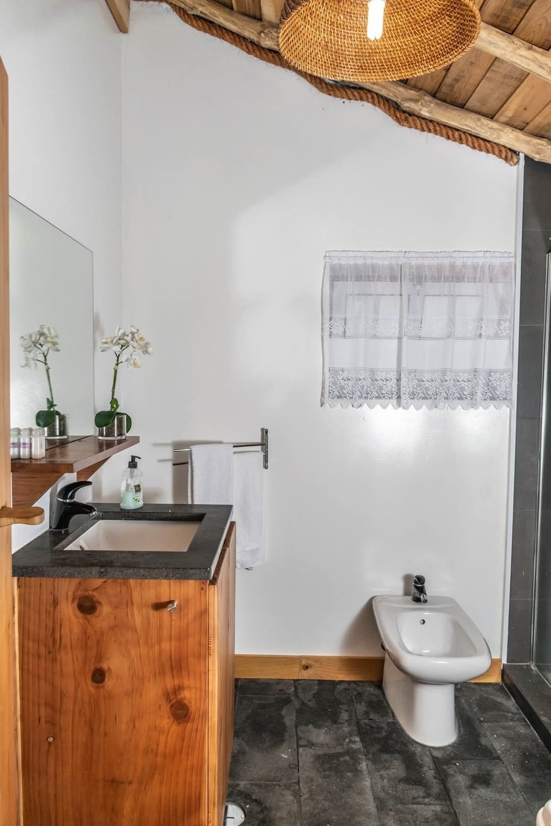 Une salle de bain avec un meuble-lavabo en bois, un lavabo, un bidet et une fenêtre ornée de rideaux en dentelle.