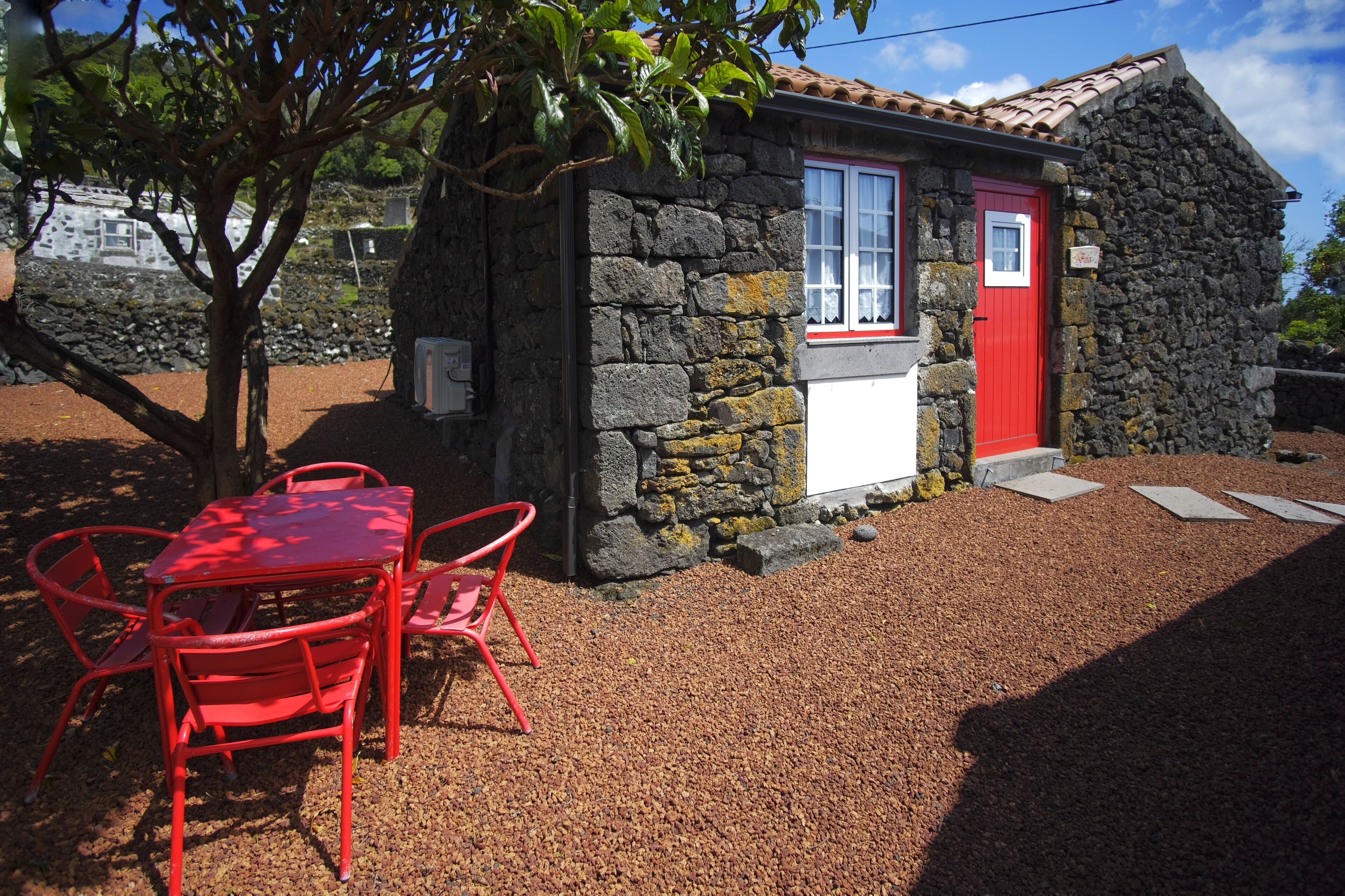 Una cabaña de piedra con puerta y ventana rojas, una pequeña mesa con sillas rojas y un camino de grava.
