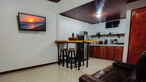 A living room with a brown leather sofa, a dining area with a wooden table and stools, a kitchen with wooden cabinets, and a framed picture of a sunset on the wall.